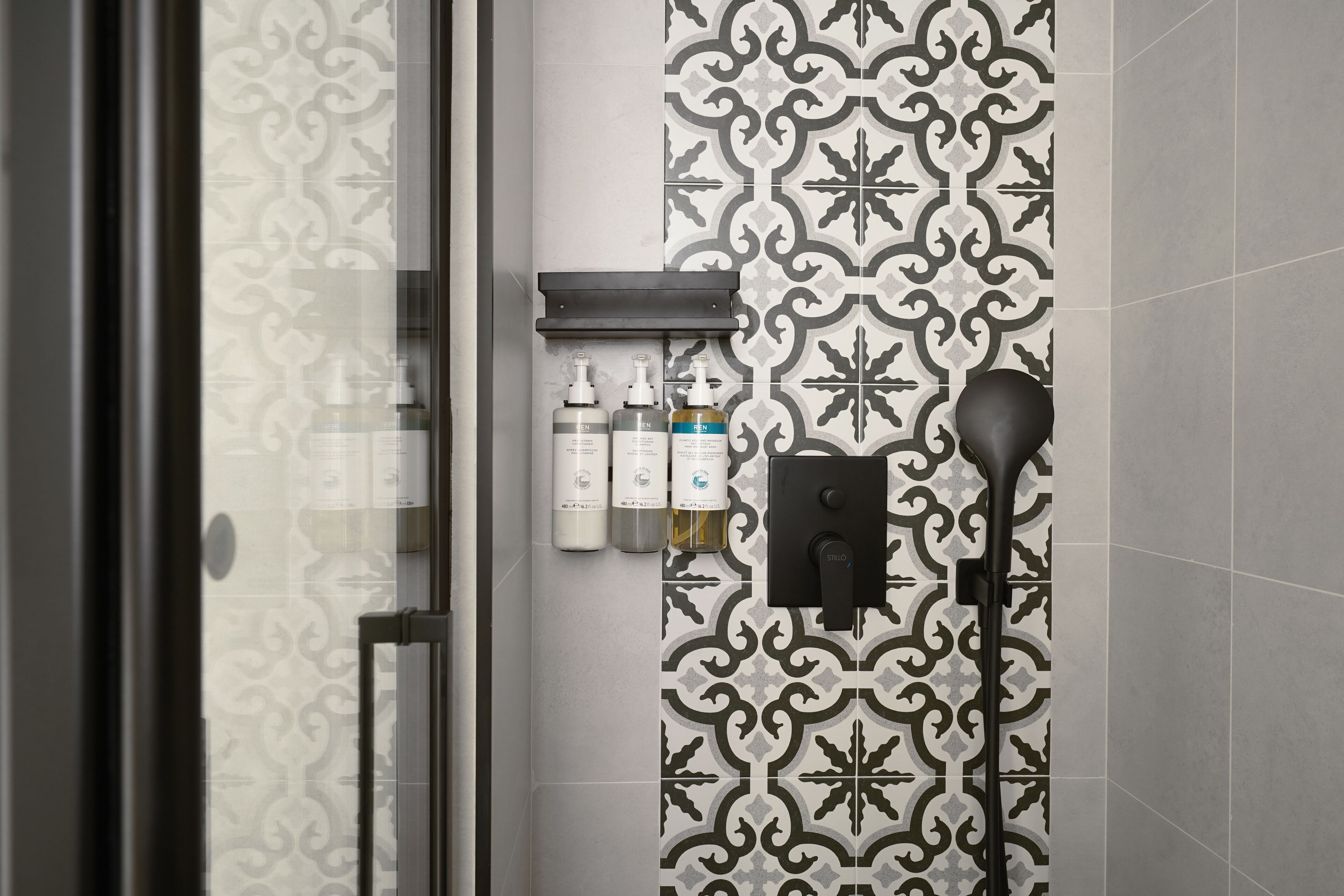 A bathroom with patterned tiles, a glass shower door, and a shelf with toiletries.