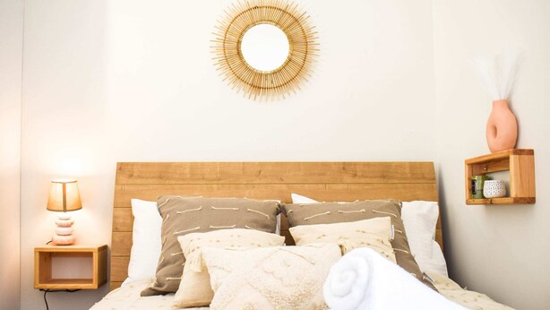 A neatly made bed with a wooden headboard, multiple pillows, a bedside lamp, a wall-mounted mirror, and a small shelf with a vase and decorative items. - Acropolis Rooftop Monastiraki. (Athina)