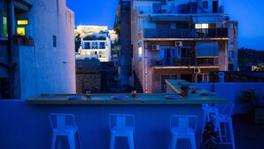 A terrace with a bar area, white bar stools, and a view of buildings in the background. - Acropolis Rooftop Monastiraki. (Athina)