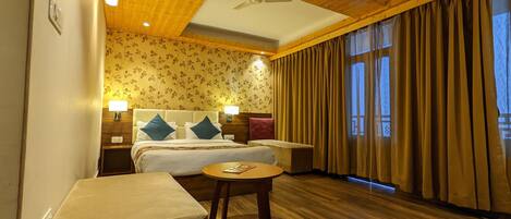 A hotel room with a bed, two bedside tables, a round table, a ceiling fan, and a window with curtains.