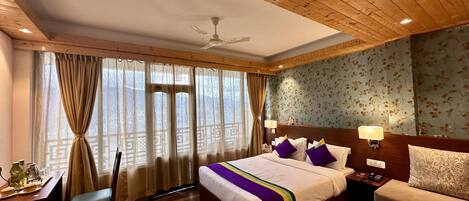 A hotel room with a wooden ceiling, a bed with a white and purple bedspread, a wooden floor, a desk with a chair, a nightstand with a lamp, and a sofa.