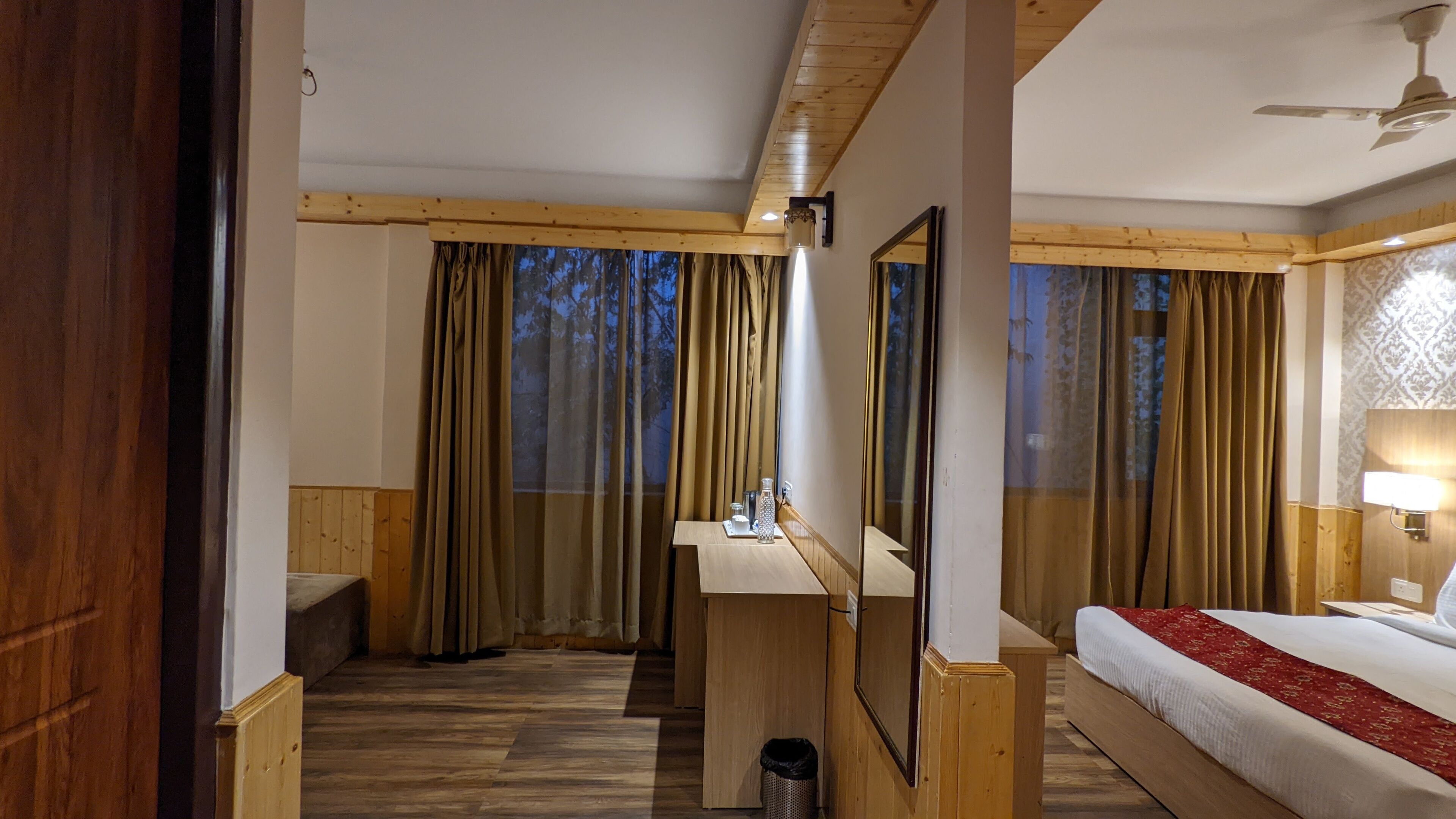 Une chambre d’hôtel avec un plancher en bois, un lit recouvert d’une courtepointe rouge, un bureau avec un miroir et une fenêtre avec des rideaux.