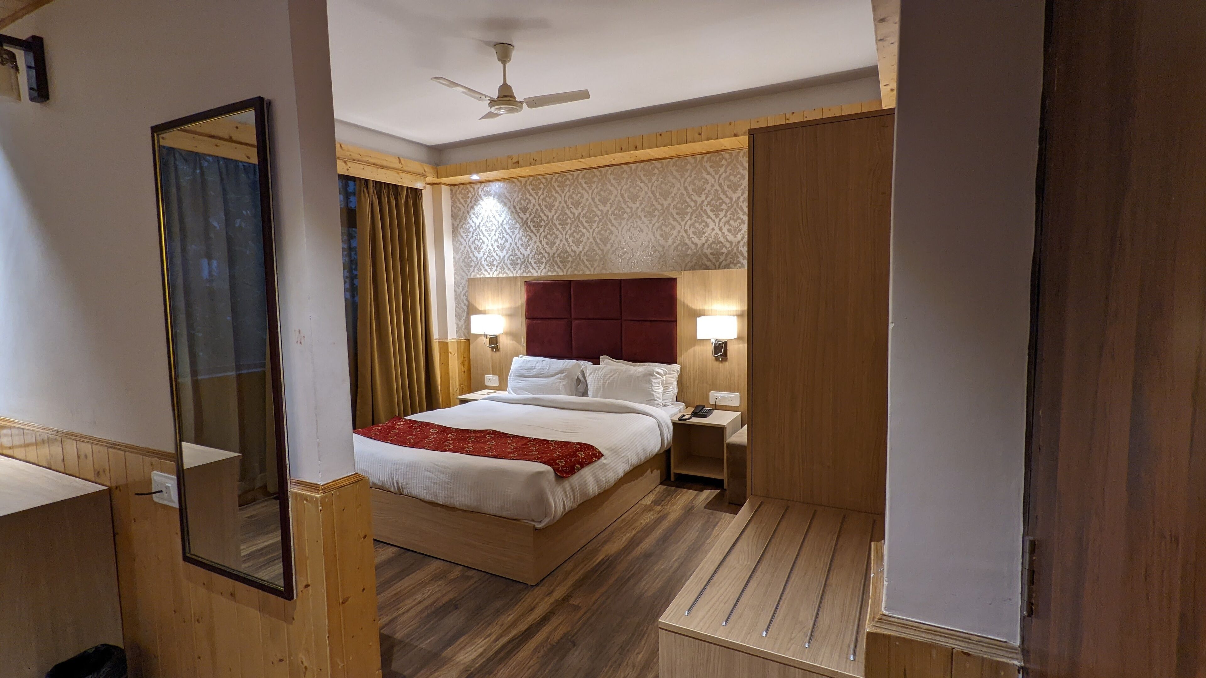 A hotel room with a wooden floor, a bed with white linens and a red throw, a wooden headboard, two bedside lamps, a nightstand, a ceiling fan, and a large mirror.