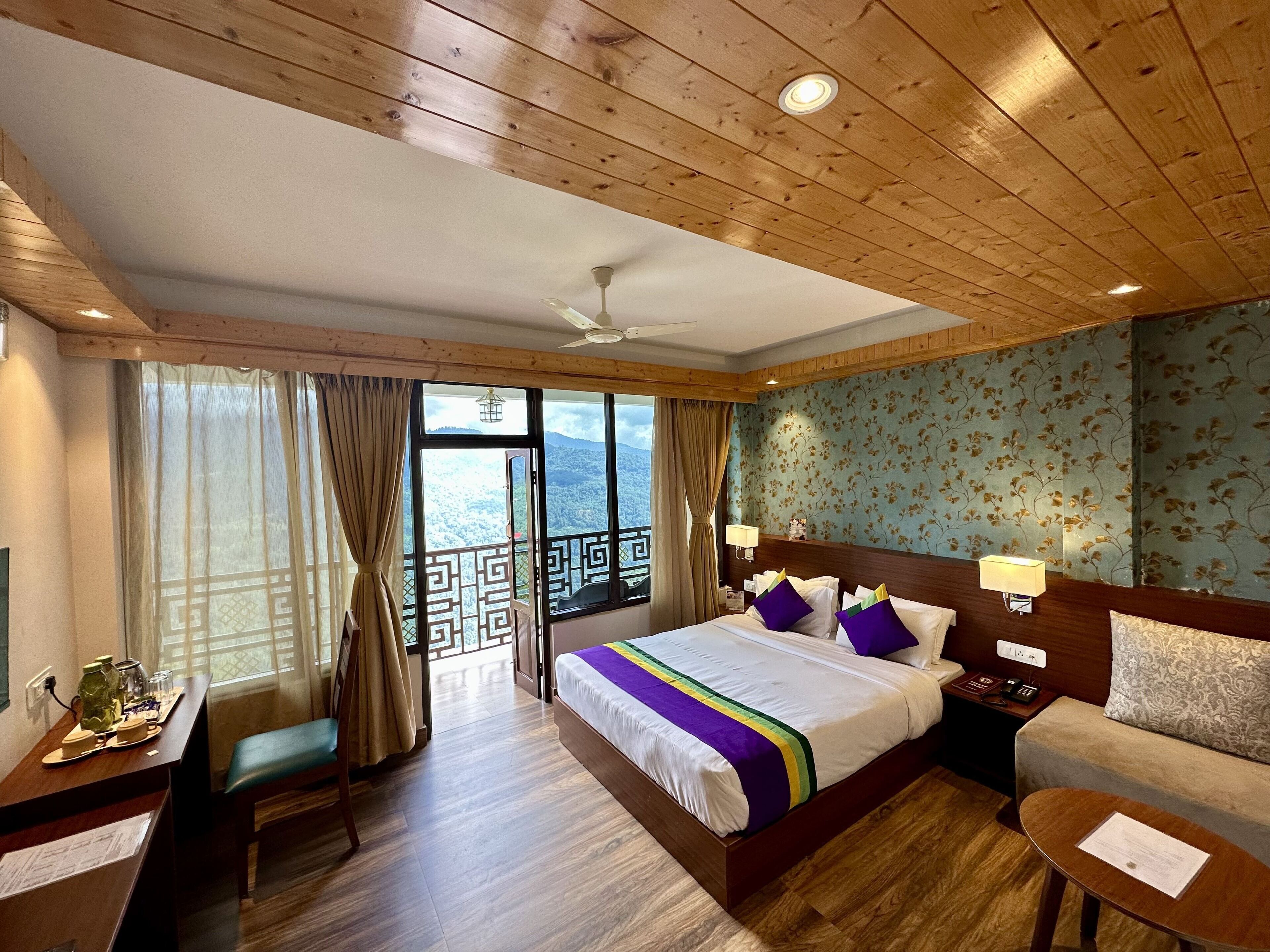 Habitación de hotel con techo de madera, una cama grande con colcha de colores, un sofá, un escritorio con una silla, y vistas a las montañas a través de la ventana.