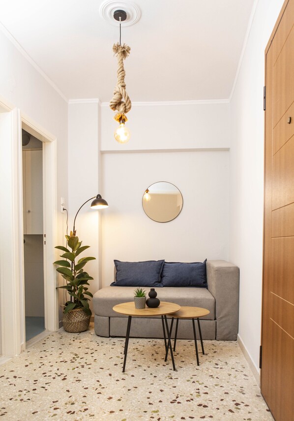 Un espace de vie compact comprenant un canapé gris, un miroir rond, un luminaire suspendu et une petite table sur laquelle est posée une plante en pot.