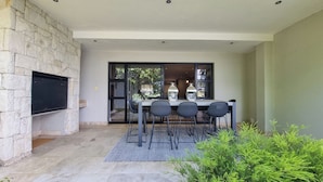 A patio with a stone wall, a flat-screen TV, a table with chairs, and a view through glass doors. - Pearl Valley Golf Lodge - Loadshedding f (Paarl)