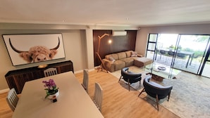 A modern living room with a sectional sofa, a dining table with chairs, a TV mounted on the wall, and a large window providing a view of the outdoors. - Pearl Valley Golf Lodge - Loadshedding f (Paarl)