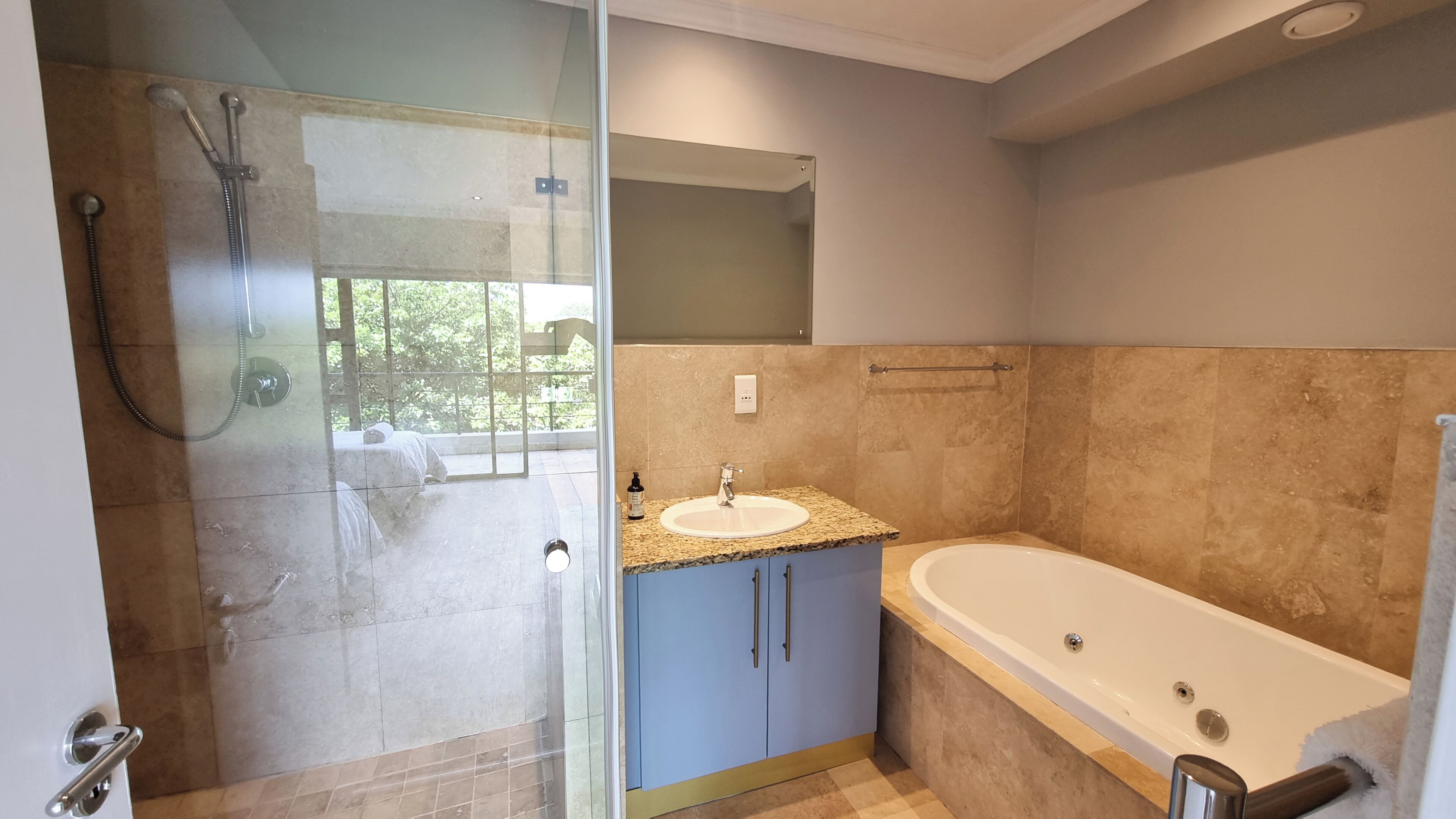 A bathroom with a glass shower enclosure, a sink, a bathtub, and a view of greenery outside.
