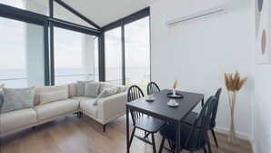 A modern living room with a sofa, dining table, and chairs, large windows with a view of the sea, and an air conditioning unit on the wall. - Mehmetbey Çaka (Persembe)