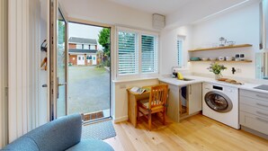 A modern kitchen with a built-in washing machine, a dining area with a table and chairs, and a large sliding glass door leading to a garden. - The Conifer Studio | Oxford Holiday Let | 1 Bed & Sleeps 1 (Oxford)