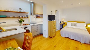 A modern bedroom with a bed, bedside table, and a TV. There is a kitchen area with a sink, cabinets, and a wooden chair. - The Conifer Studio | Oxford Holiday Let | 1 Bed & Sleeps 1 (Oxford)