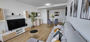 Ein modernes Wohnzimmer mit einem Flachbildfernseher, einem grauen Sofa, einem Couchtisch und einem Essbereich mit Stühlen und einem Tisch.