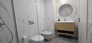 A modern bathroom with a marble shower, a toilet, a bidet, and a wooden vanity with a round mirror.