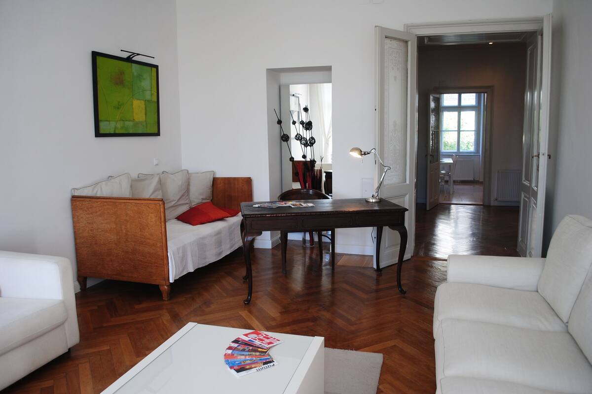 A living room with a white sofa, a wooden coffee table, and a dark wooden side table with a lamp. There is a painting on the wall and a door leading to another room.