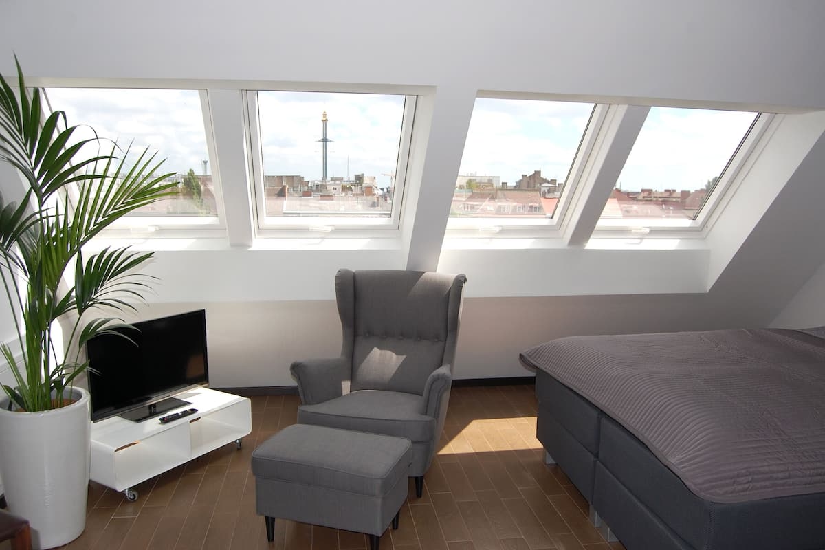 A modern loft bedroom with a large skylight, a grey armchair, a TV on a white console, and a grey bed.