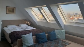 A bedroom with a bed, a nightstand, a chair, and a window with a city view. - Kunsthaus Apartments (Vienna)