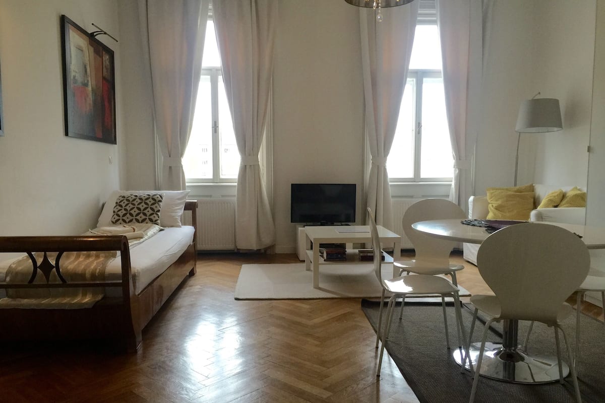 A modern living room with a wooden floor, a sofa, a dining table with chairs, a TV, and a window with curtains.