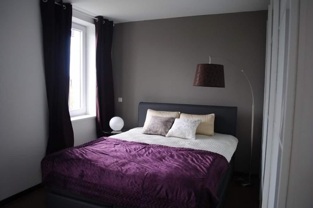 A bedroom with a bed, dark curtains, a bedside lamp, and a window.