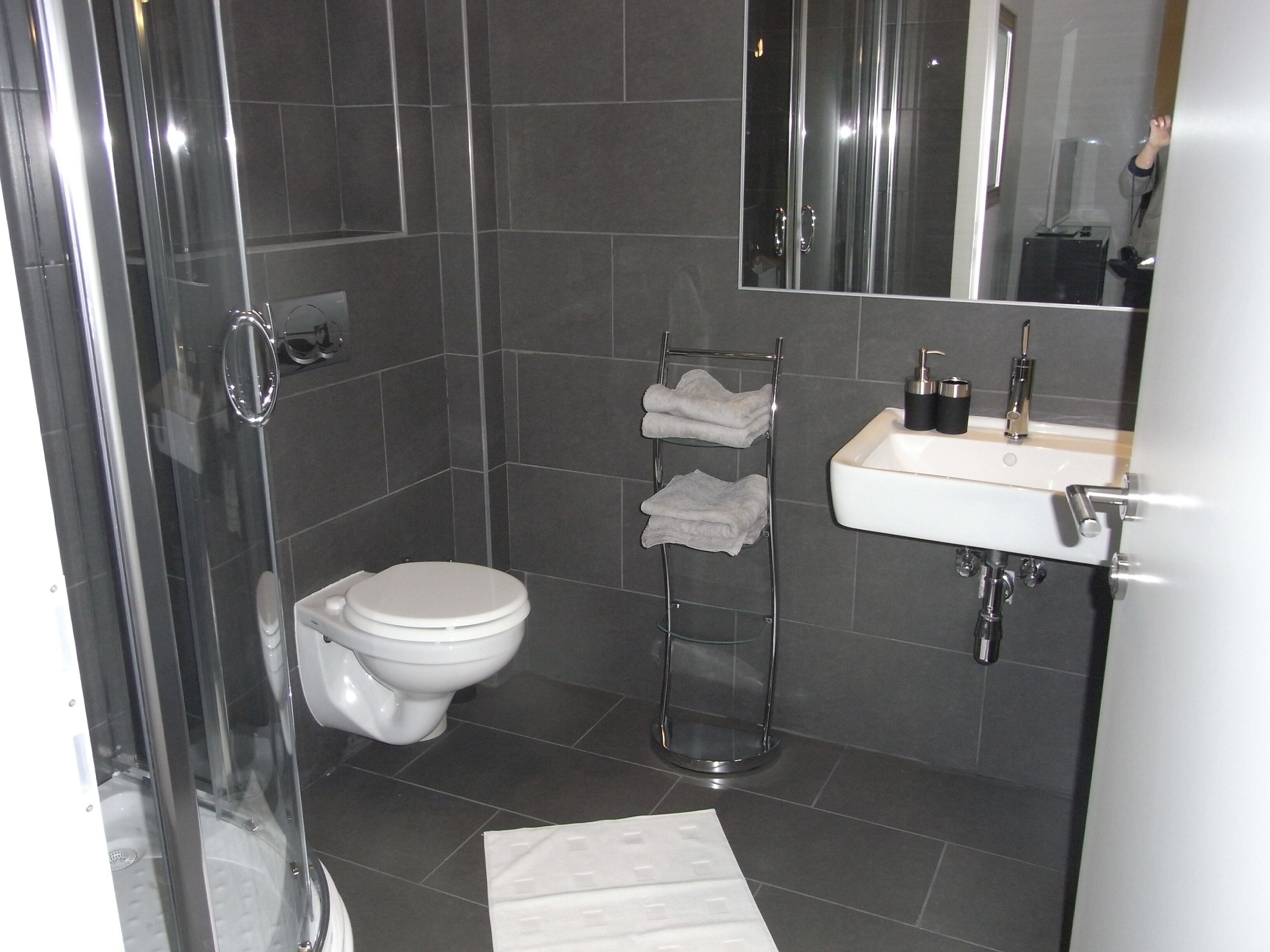 A modern bathroom with a toilet, a glass shower enclosure, a towel rack with folded towels, and a sink with a mirror above it.