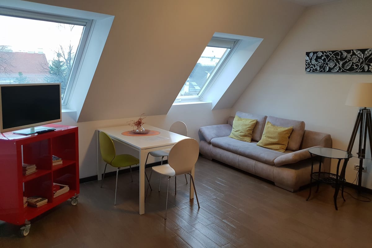 A small, modern living room with a loft-style ceiling, a red bookshelf, a white desk, a green chair, a beige sofa with yellow cushions, and a small side table.