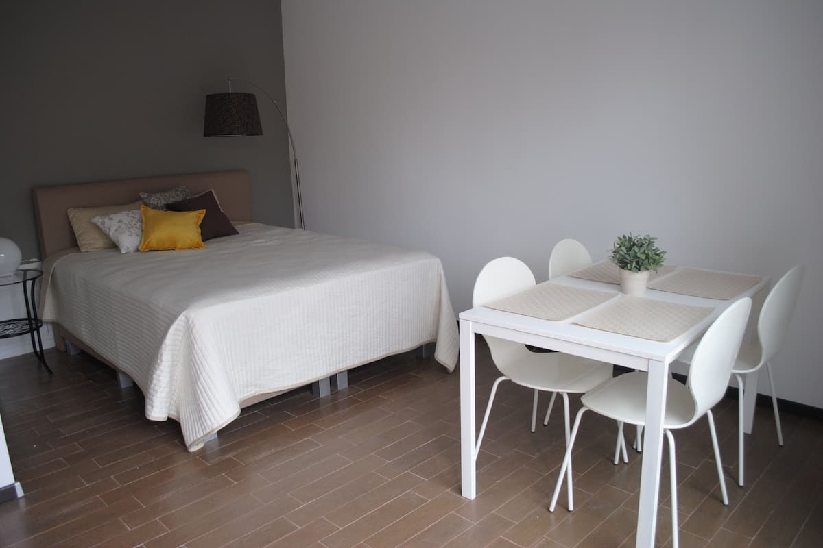 A bedroom with a bed, bedside table, lamp, and a dining area with chairs and a table.