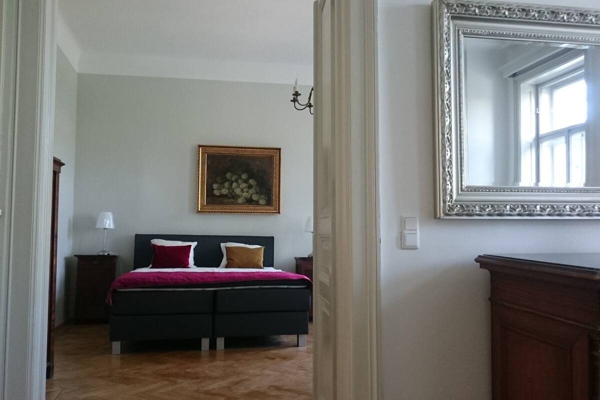 A bedroom with a bed, a painting, a mirror, and a dresser.