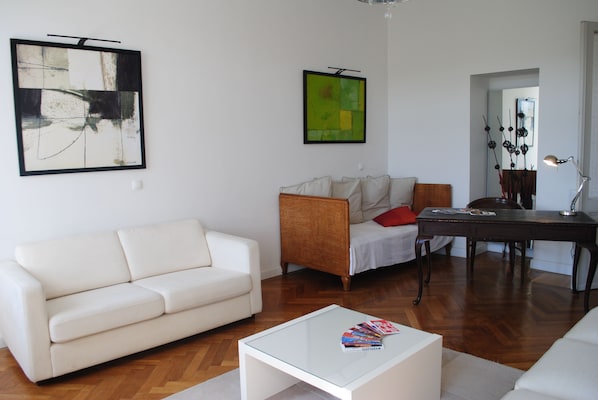 Ein Wohnzimmer mit einem weißen Sofa, einem Glastisch, einem Holznachttisch und zwei Bildern an der Wand.
