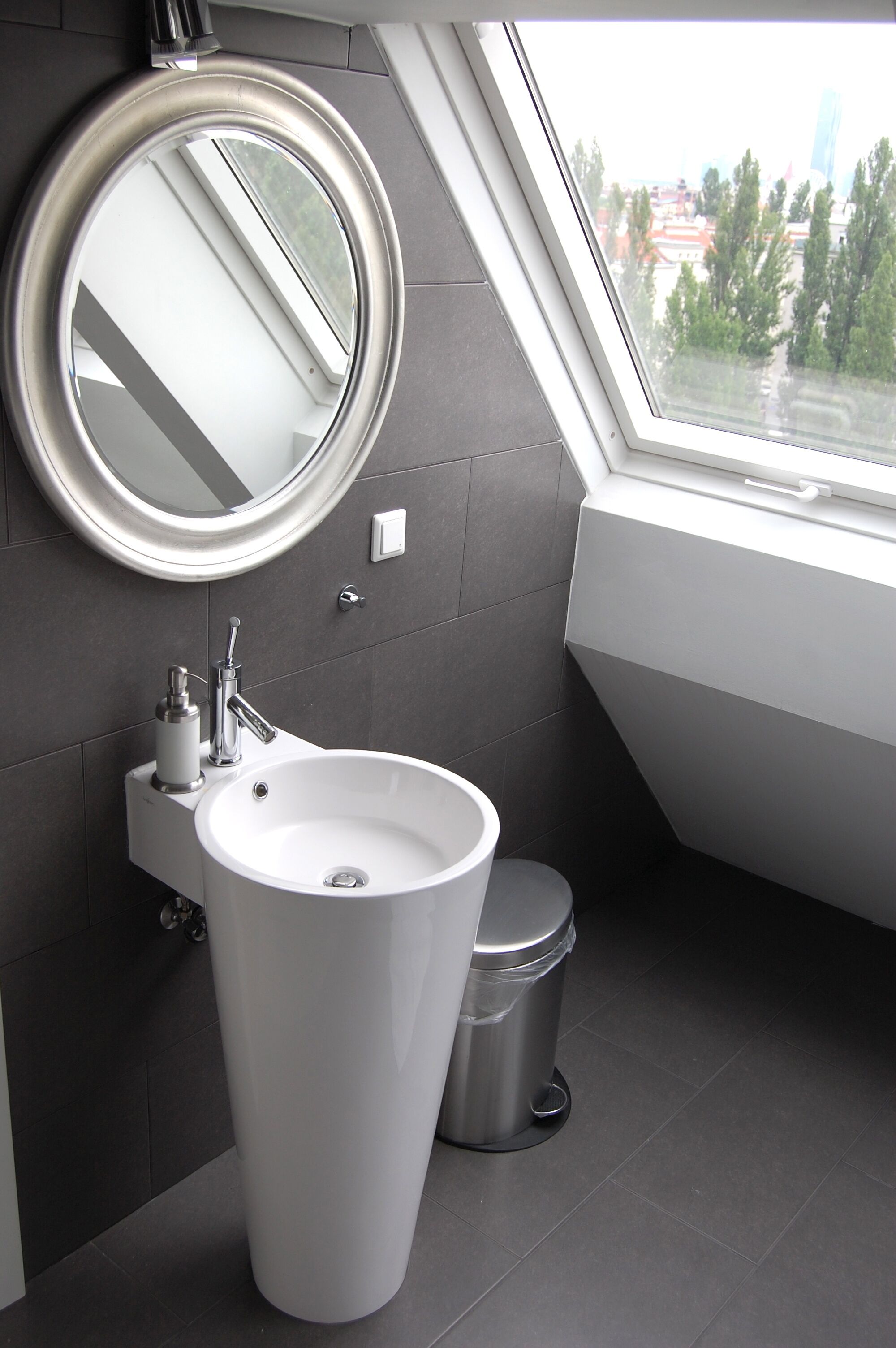 A modern bathroom with a large sink, a round mirror, and a window providing a view of the city.