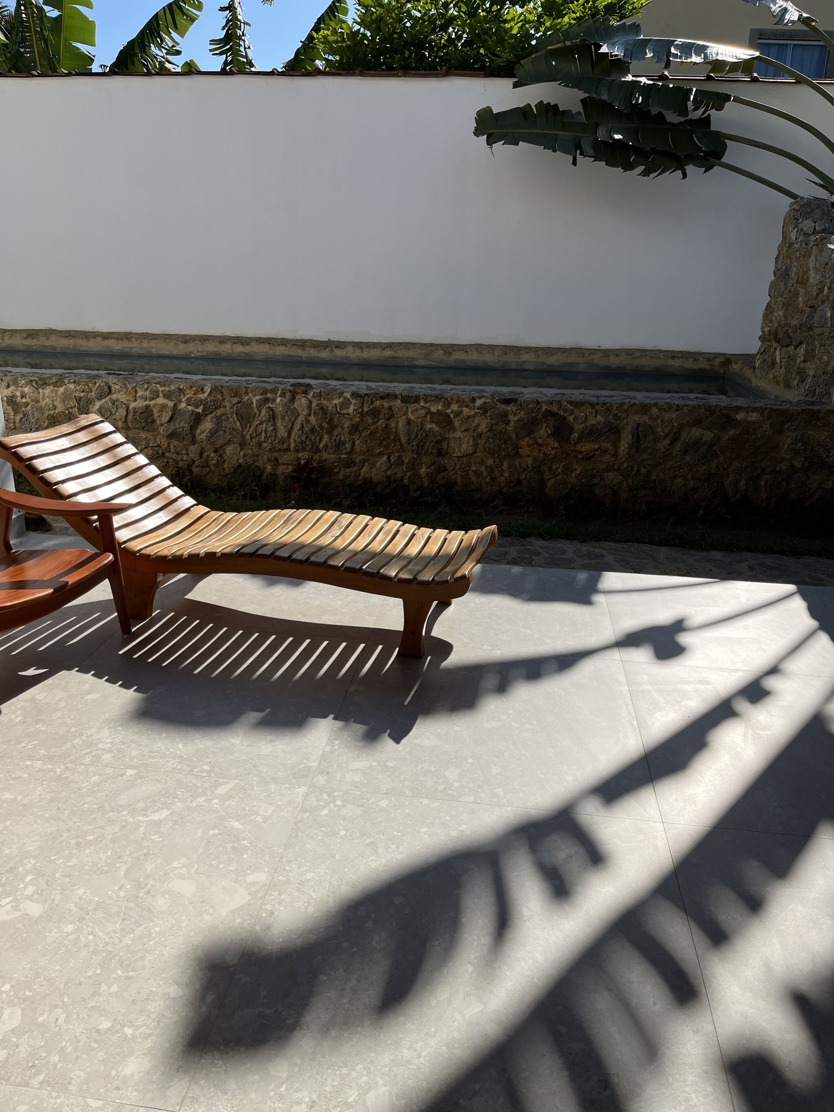 A wooden lounge chair on a tiled patio with a white wall and stone steps in the background.