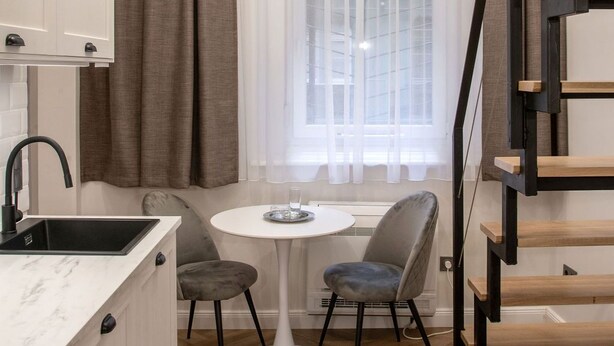 Eine kleine Küche mit weißer Arbeitsplatte, einem schwarzen Spülbecken und einem Essbereich mit zwei Stühlen und einem weißen Tisch. Ein Fenster mit transparenten Vorhängen ist sichtbar, und eine Treppe mit Holzwand ist rechts zu sehen.