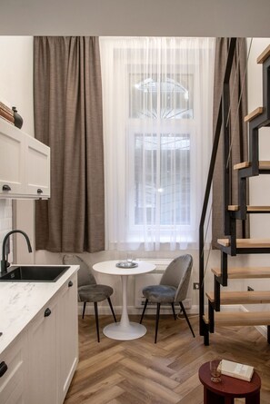 Une cuisine compacte avec un plan de travail blanc, un évier noir et un petit coin repas composé de deux chaises et d’une table blanche. Une fenêtre avec des rideaux légers est visible, et un escalier avec une rampe en bois se trouve sur la droite.