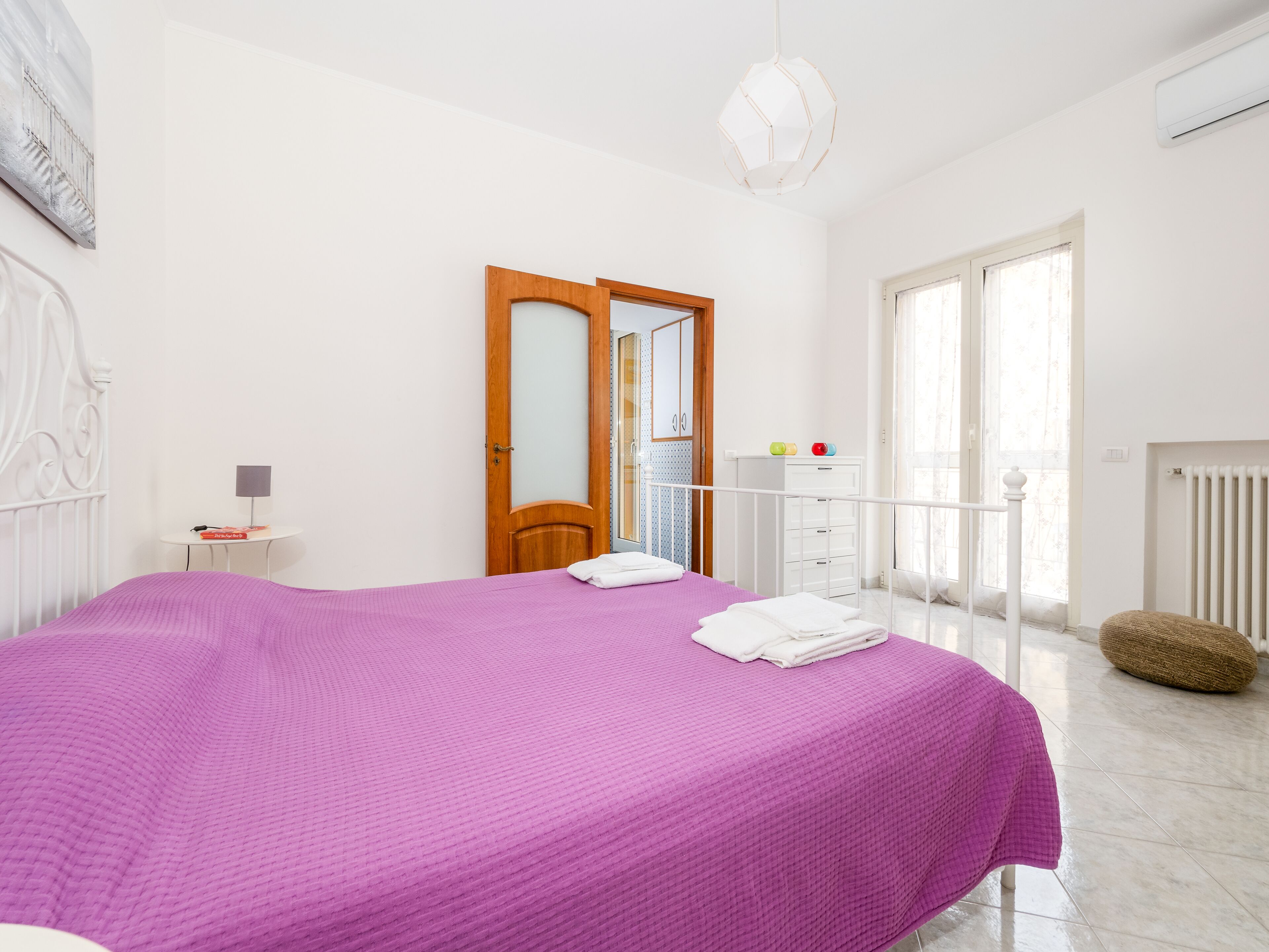 A bedroom with a large bed, a wooden door, a white radiator, and a window with curtains.