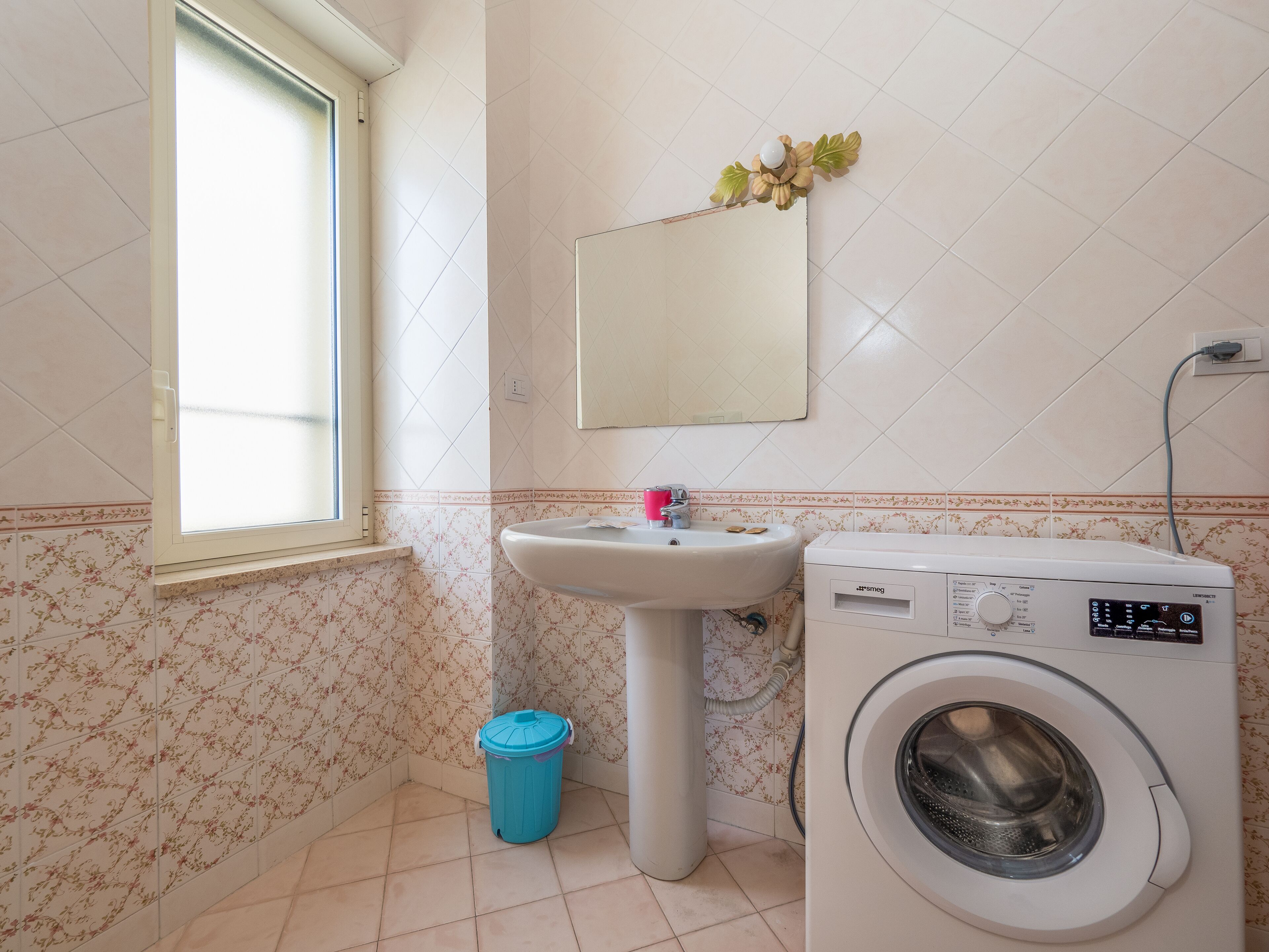 Une salle de bain avec une fenêtre, un lavabo, un lave-linge et une poubelle.