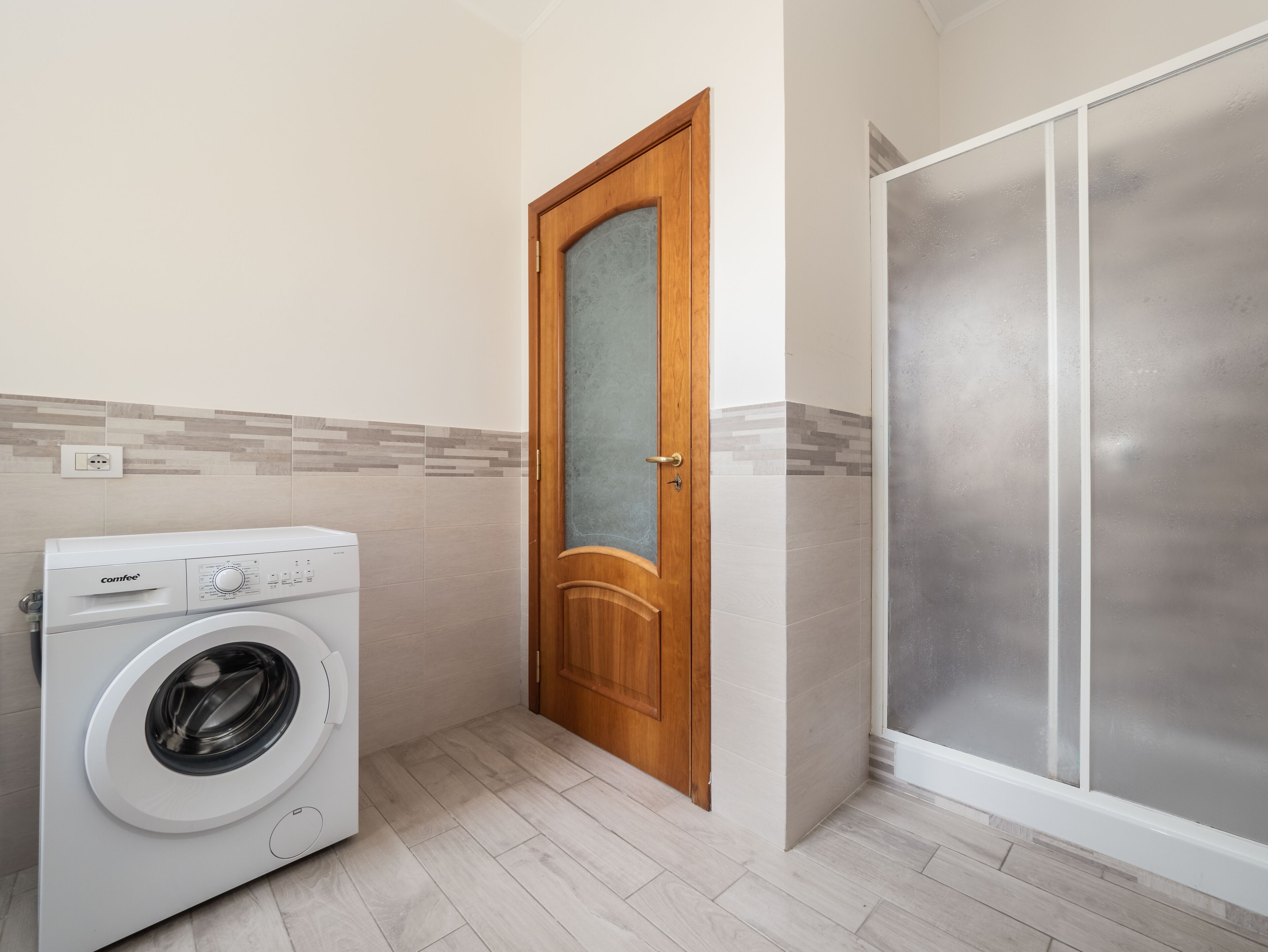 A compact laundry room with a washing machine, a wooden door, and a frosted glass shower enclosure.