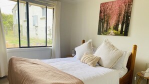 Een slaapkamer met een groot raam, een bed met witte beddengoed, een houten hoofdbord, en een schilderij van bomen aan de muur.