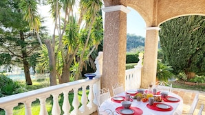 An outdoor dining area with a white table set for four, surrounded by white balustrades and columns, overlooking a lush garden and a pool.