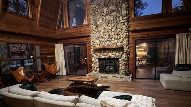 A spacious living room with a stone fireplace, wooden beams, and large windows. - Welcome To Orlando's Chic Cabin! KING BED, FIREPLACE, SAUNA, HOT TUB, GAMES  (Sorrento)