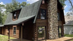 A log cabin with a stone chimney, large windows, and a wooden deck. - Welcome To Orlando's Chic Cabin! KING BED, FIREPLACE, SAUNA, HOT TUB, GAMES  (Sorrento)
