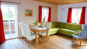 Um quarto com uma mesa de jantar de madeira, quatro cadeiras, um sofá verde e uma poltrona azul. Há duas janelas com cortinas vermelhas e um quadro na parede.