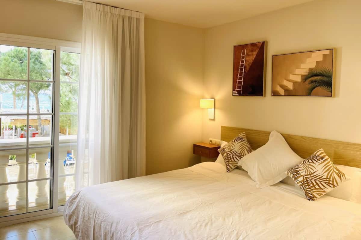 A hotel room with a large bed, a nightstand, a lamp, and a view of the outdoors through a sliding glass door.