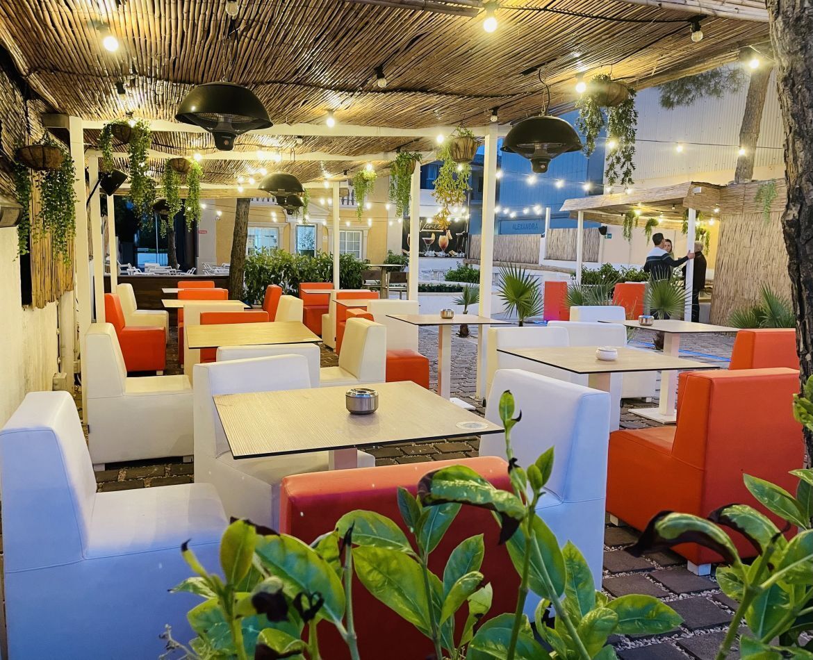 An outdoor seating area with tables and chairs, surrounded by greenery and a thatched roof.