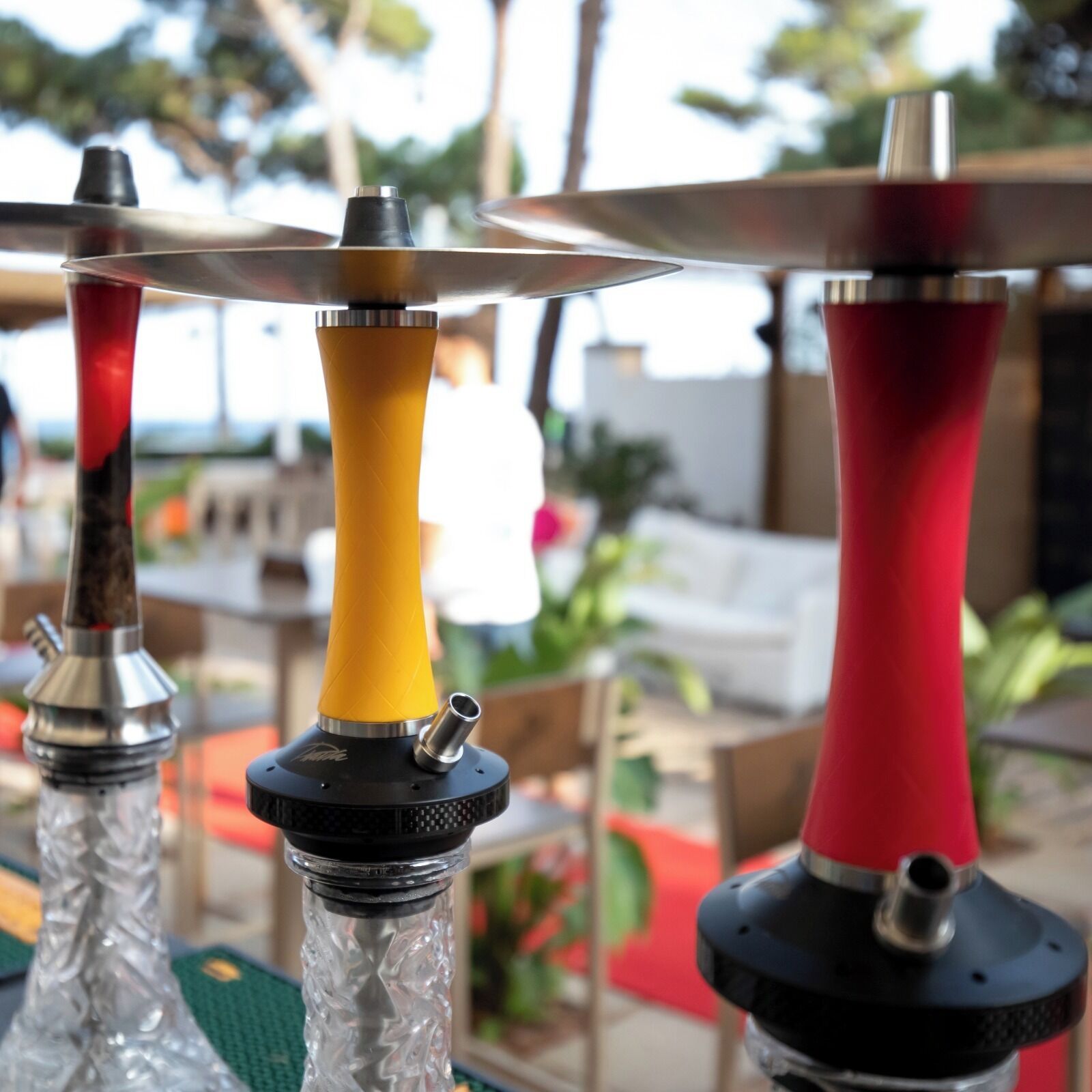 Une vue rapprochée de trois narguilés aux bases de couleurs différentes, placés sur un fond extérieur flou, avec de la verdure et du mobilier.