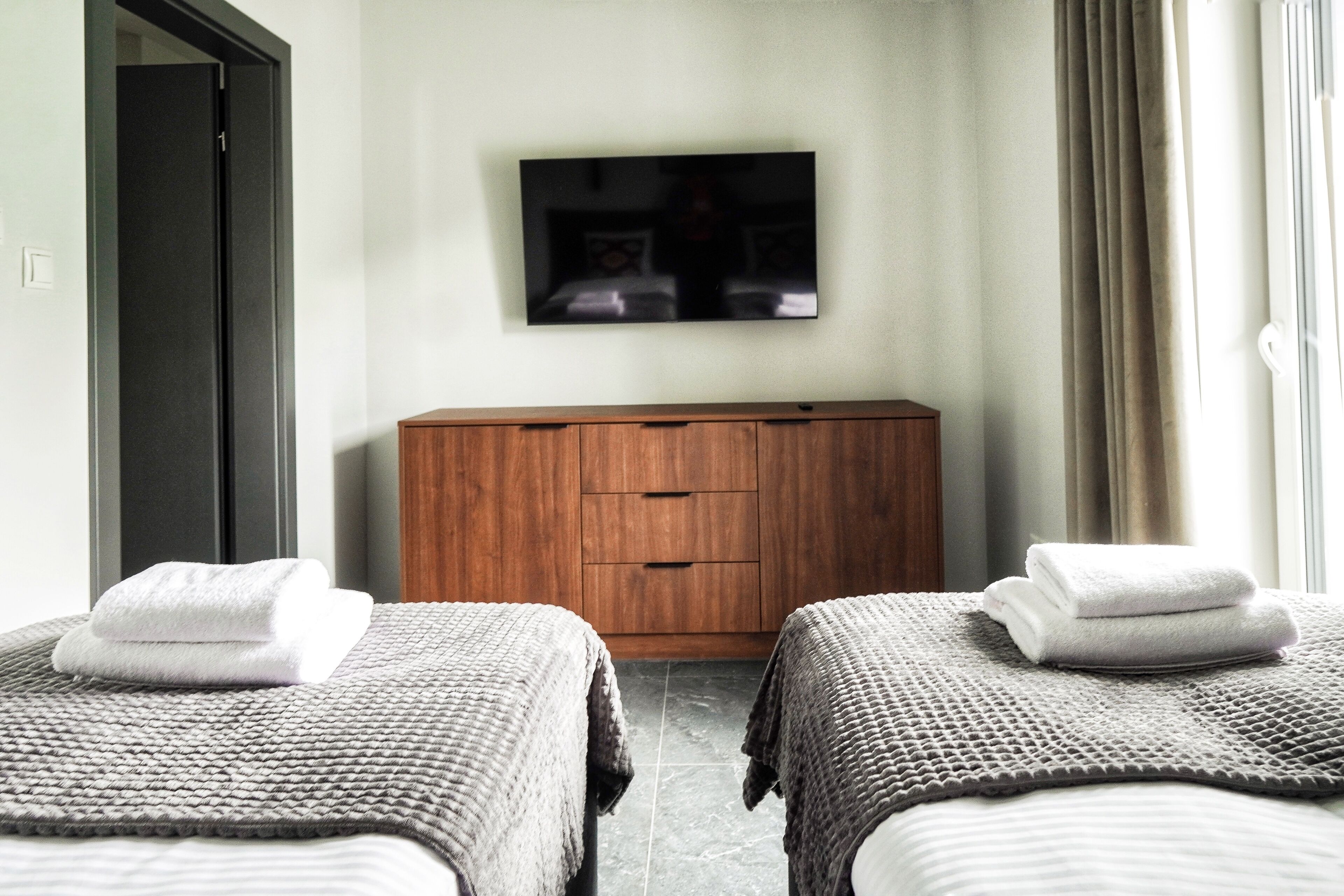 Ein Hotelzimmer mit zwei Betten, einer Holzkommode, einem Flachbildfernseher an der Wand und einem Fenster mit Vorhängen.