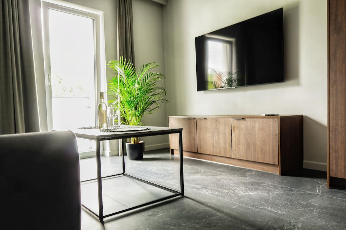Een moderne woonkamer met een salontafel met glazen blad, een potplant, een houten kast en een flatscreen televisie die aan de muur is bevestigd.