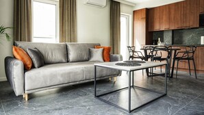Una sala de estar moderna con un sofá gris, una mesa de centro y una zona de cocina con armarios de madera y encimera de mármol.