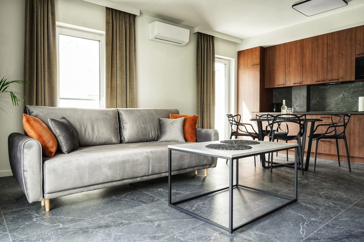 Een moderne woonkamer met een grijze bank, een salontafel en een keukenhoek met houten kasten en een marmeren aanrecht.