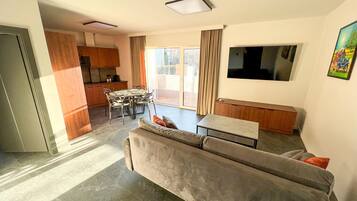 A modern living room with a grey sofa, a dining table, and a kitchen area with wooden cabinets.