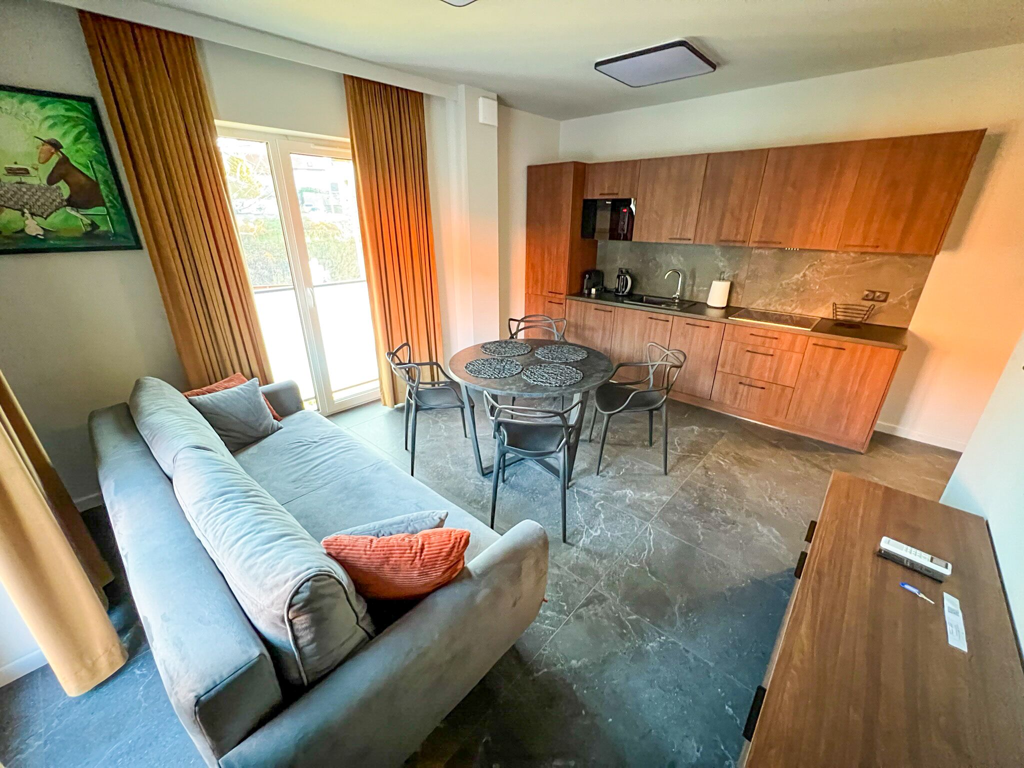 A modern living room with a grey sofa, a dining table with chairs, a kitchen area with wooden cabinets, and a large window with curtains.