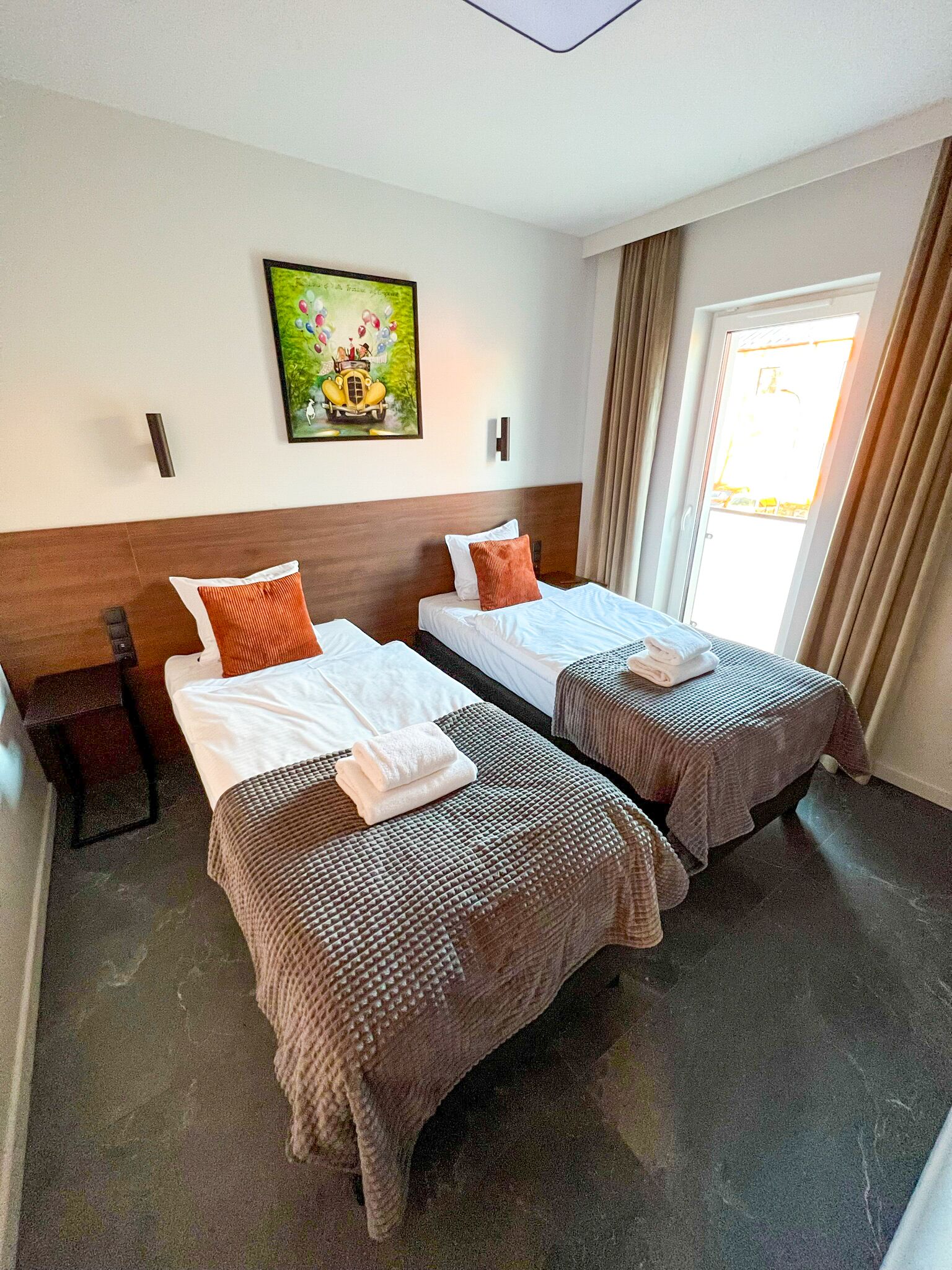 A hotel room with two beds, a painting on the wall, and a window with curtains.