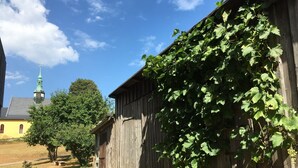 Un bâtiment rustique en bois, avec un mur couvert de vigne, un escalier en pierre et, en arrière-plan, le clocher d’une église.